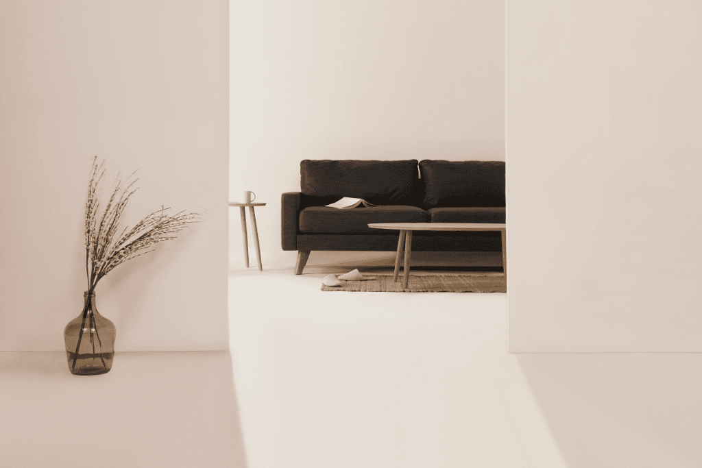 Minimalist living room with a dark grey sofa, wooden coffee table, side table with a cup, and a glass vase holding dried branches against soft neutral walls.